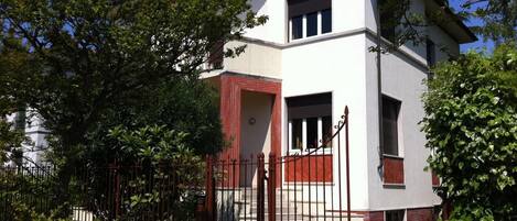 Un edificio de dos pisos con fachada blanca y detalles en rojo, que cuenta con una entrada protegida por una reja y una escalera que conduce al piso superior.