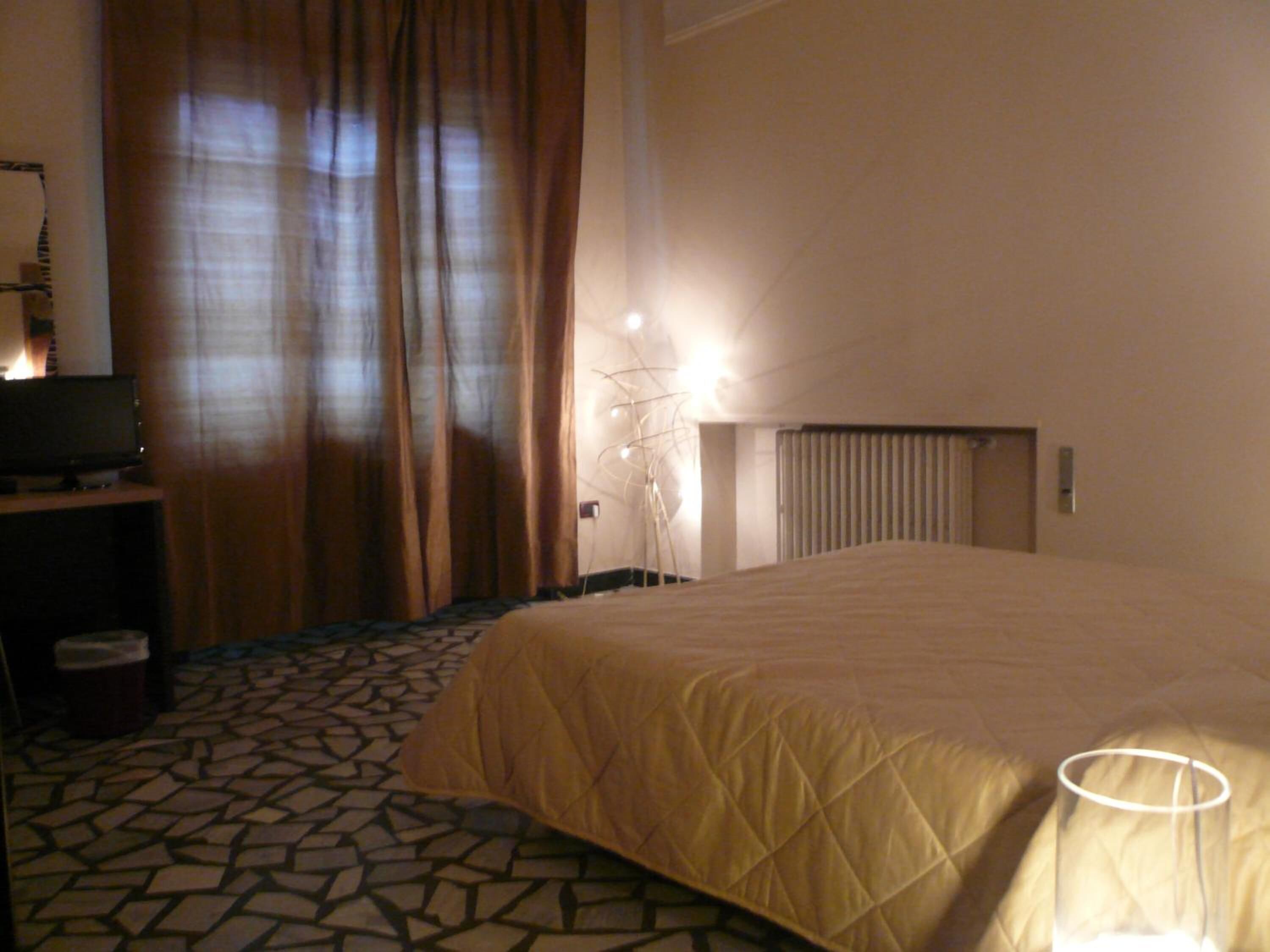 A hotel room with a bed, a desk with a television, a radiator, and a patterned floor.