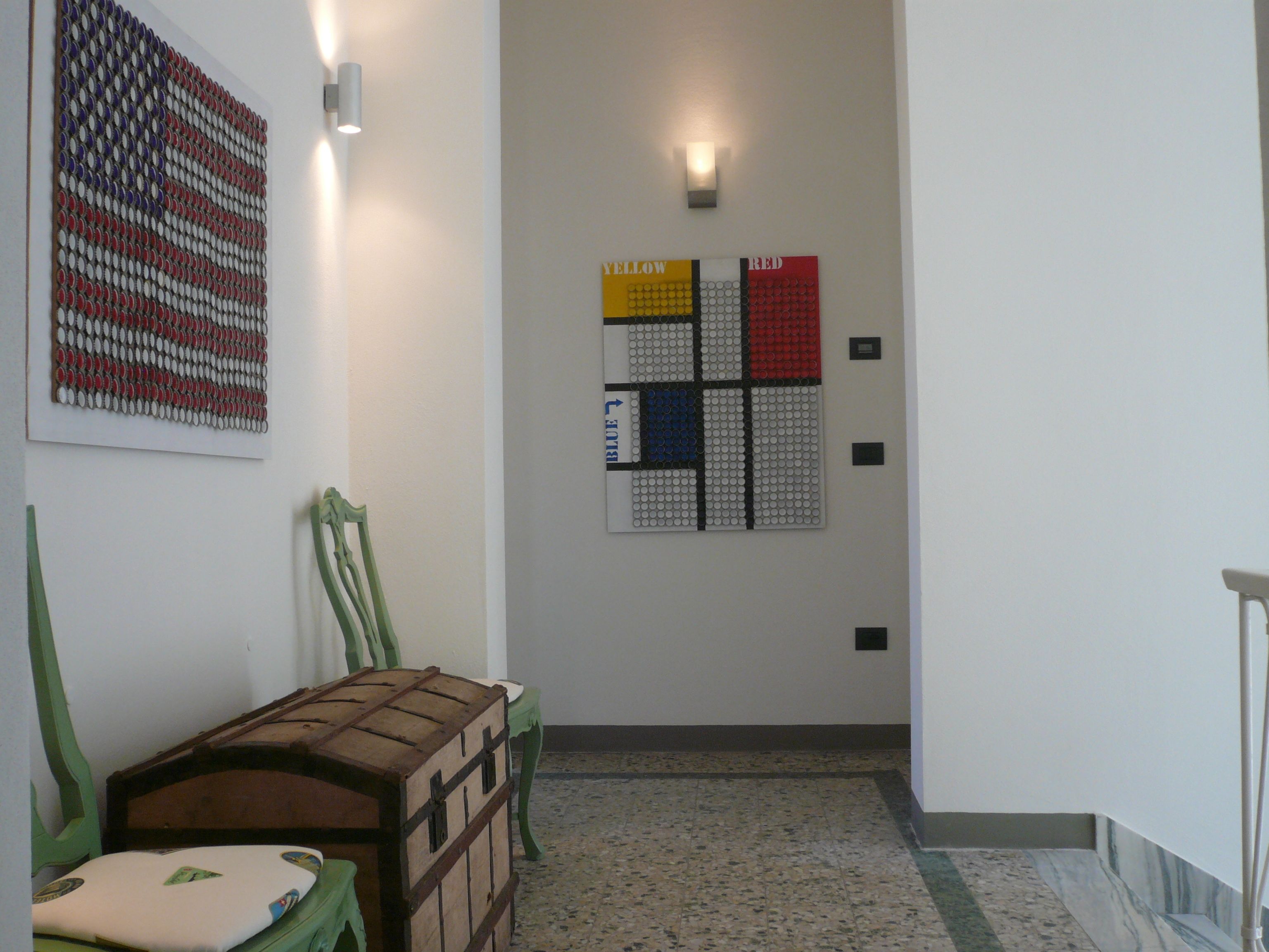 Un pasillo con un banco de madera, una silla verde y una obra de arte en la pared. Se observan dos lámparas de pared y una puerta con un letrero encima.