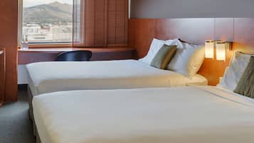 Habitación de hotel con dos camas, un escritorio y una ventana con vistas a la ciudad y montañas.