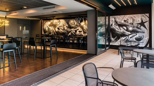 Ein modernes Restaurant-Interieur mit Tischen, Stühlen, einer Neonreklame und abstrakter Wandgestaltung.