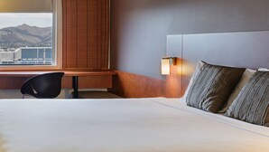 Ein Hotelzimmer mit einem großen Bett, einem Schreibtisch und einem Stuhl. Ein Fenster bietet Blick auf Berge und Gebäude.