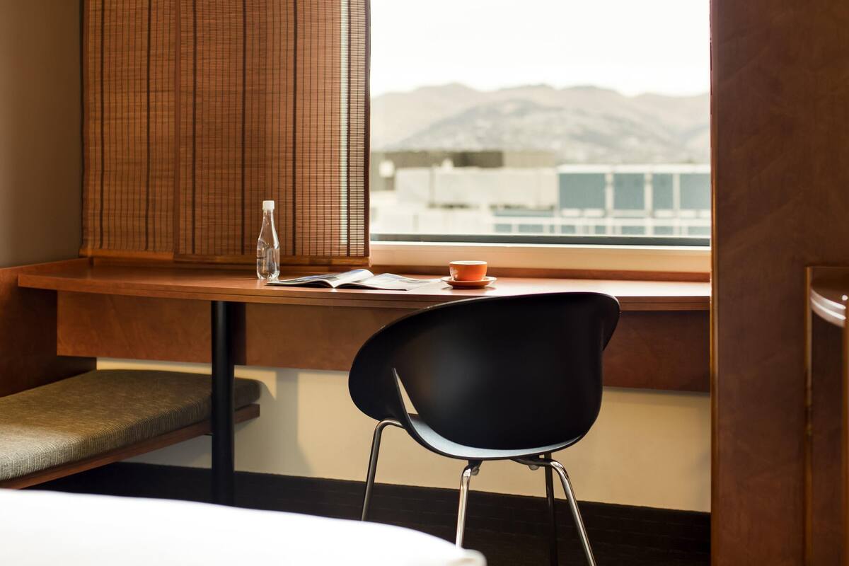 Ein Hotelzimmer mit einem Holztisch, einem schwarzen Stuhl, einer Wasserflasche und einer Tasse am Fensterbrett.