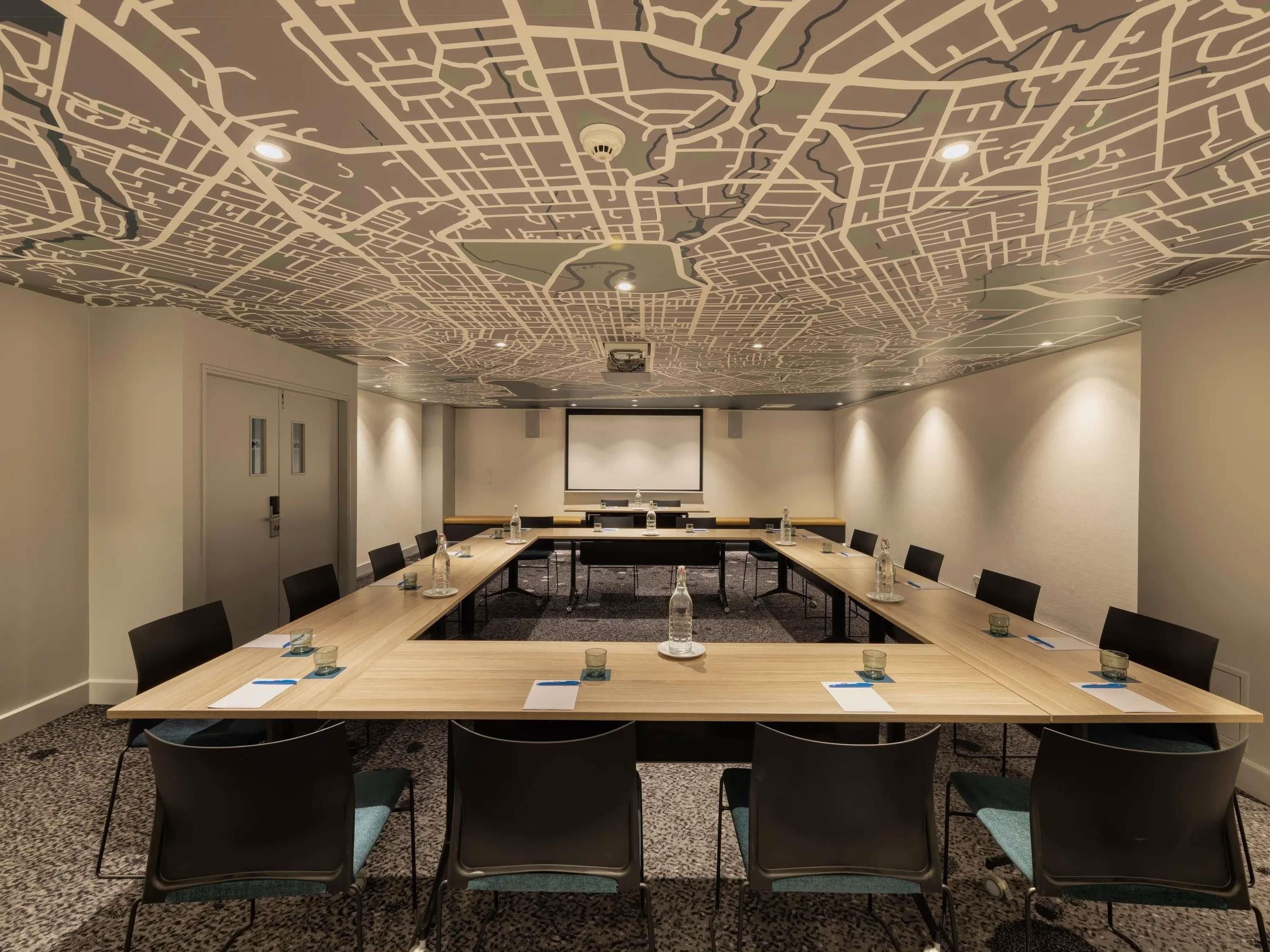A conference room with a long table, chairs, a projector screen, and a ceiling with a city map design.