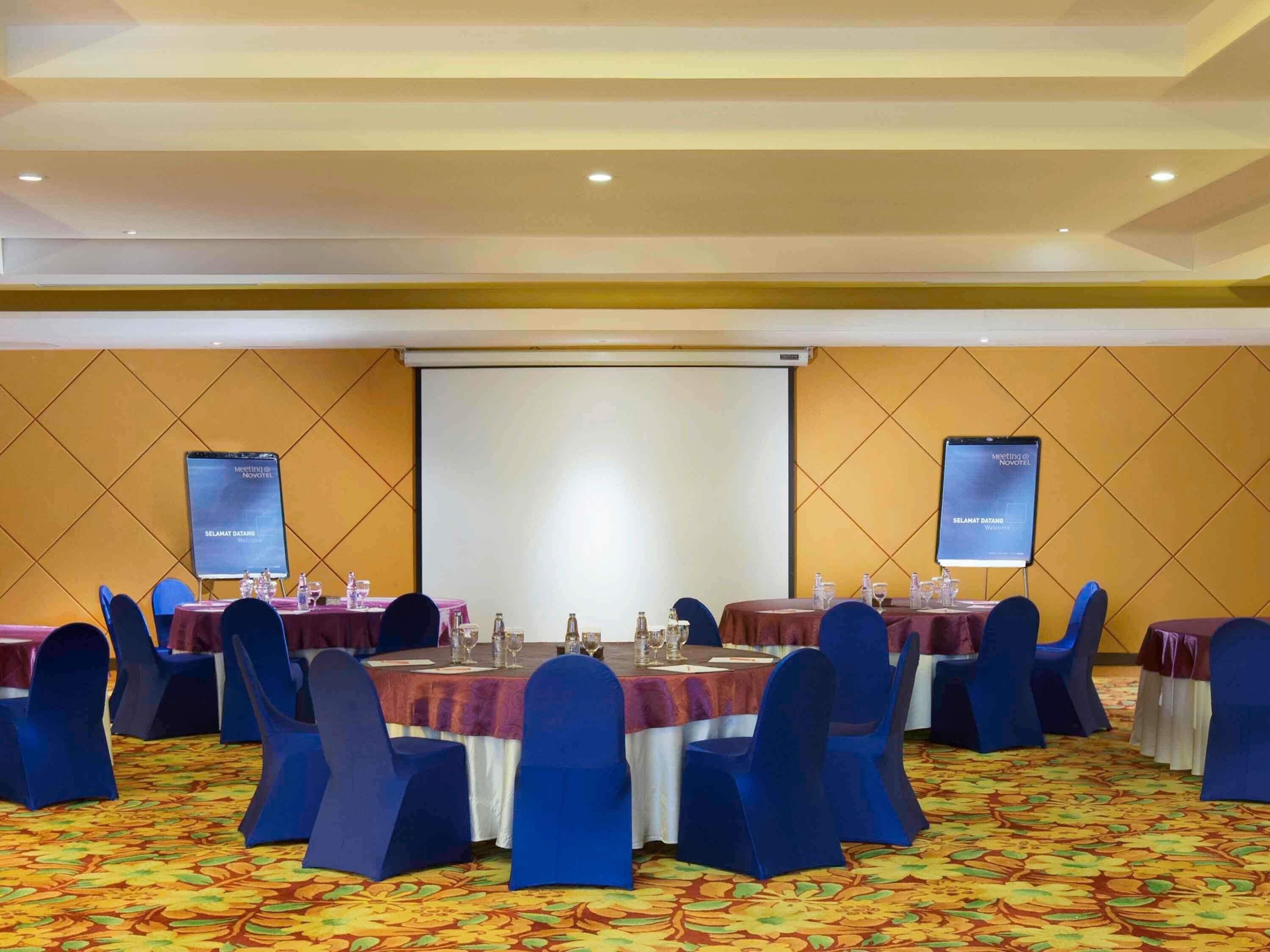 Un salón de conferencias con mesas dispuestas para reuniones, una pantalla de proyector y una pantalla de presentación.