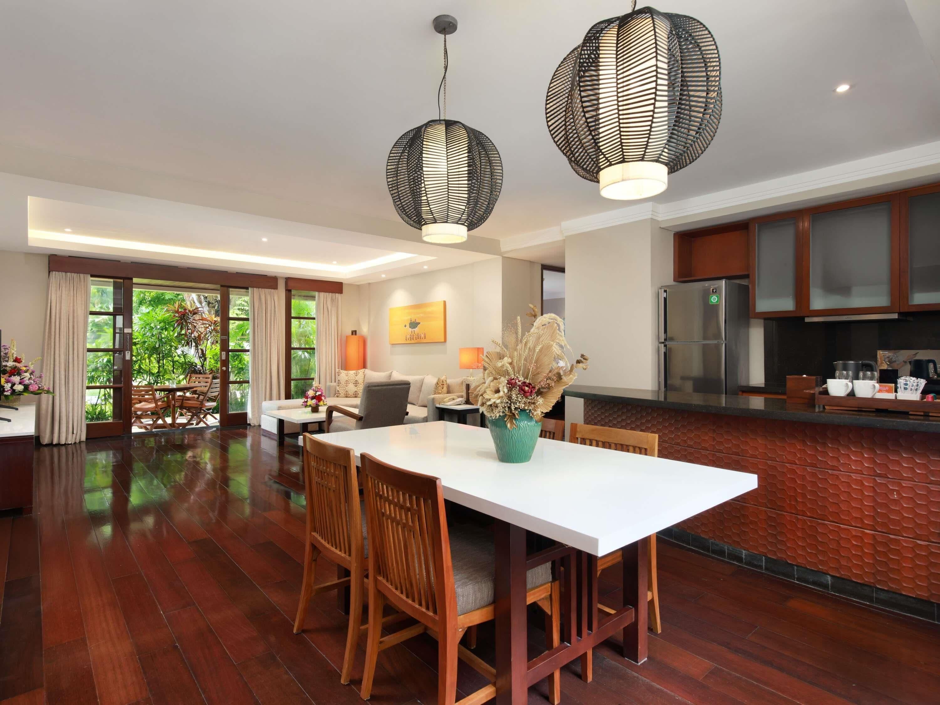 A spacious living area with a dining table, wooden chairs, and a sofa. There are two pendant lights, a vase with dried flowers, and a kitchen area with a refrigerator and cabinets.