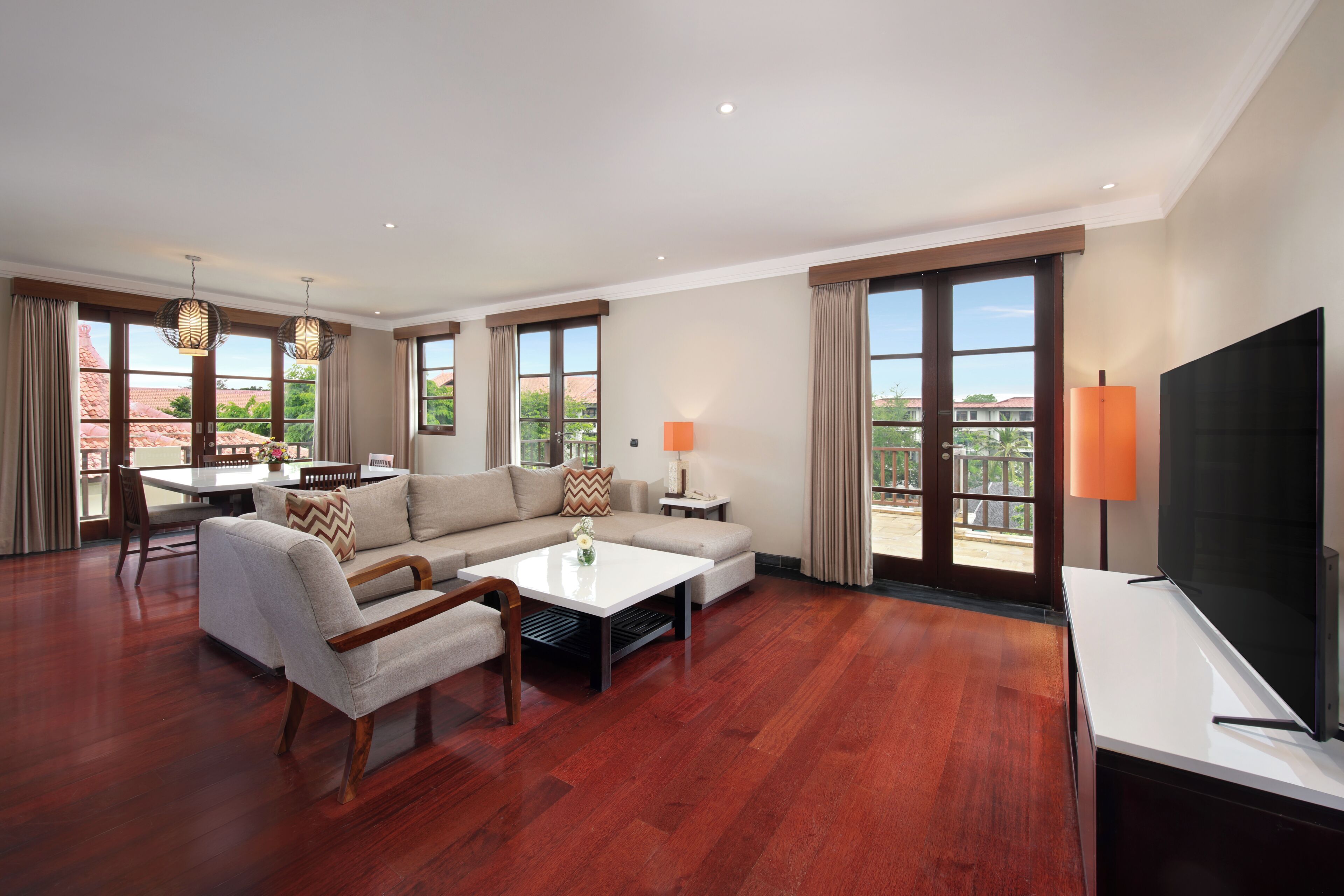 A spacious living room with a sofa, coffee table, and TV, large windows with curtains, and a balcony view.