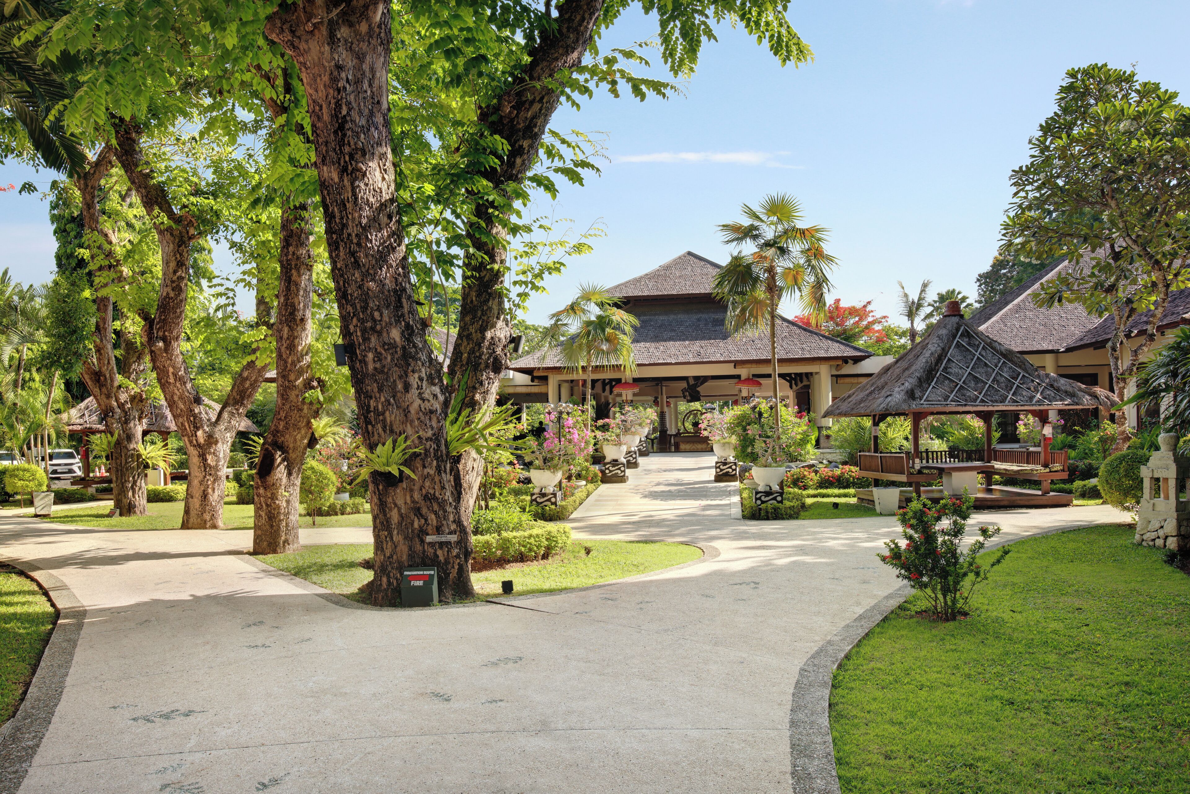 A resort with a central pathway leading to a building with a thatched roof, surrounded by lush greenery and palm trees.