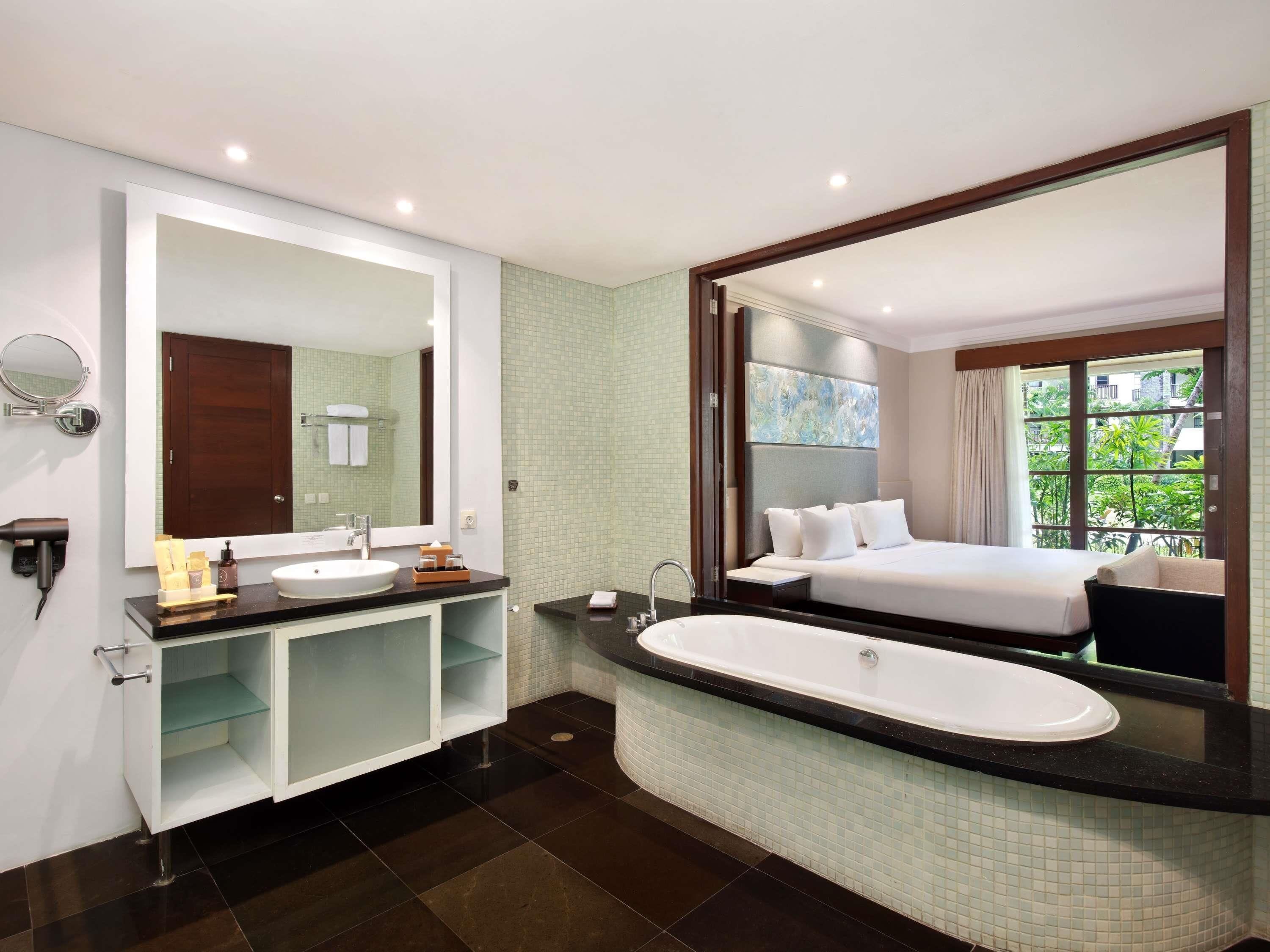 A modern bathroom with a large mirror, a sink, a bathtub, and a bed with a view of greenery outside.