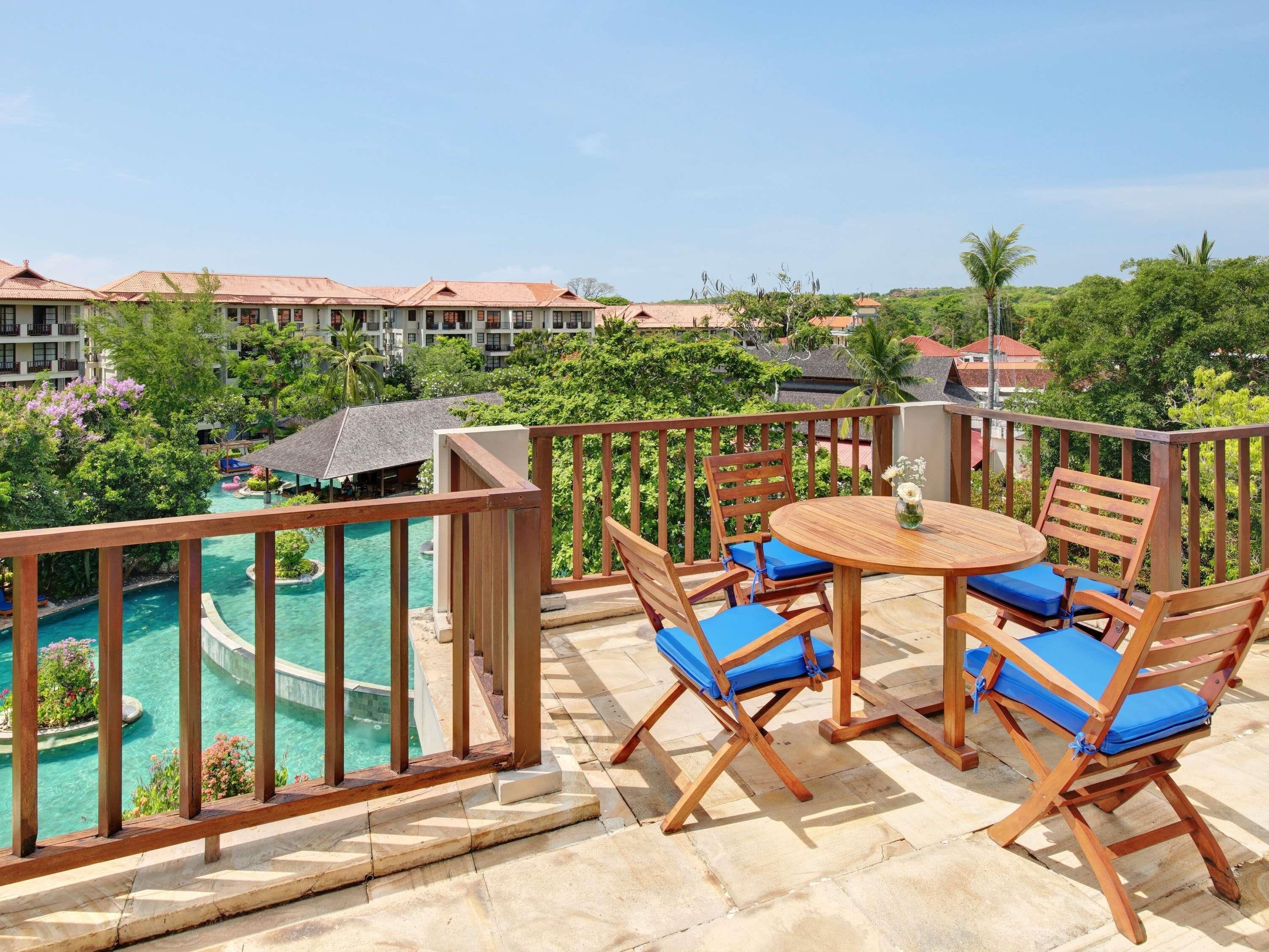 A balcony with a wooden table and chairs, overlooking a pool and lush greenery.