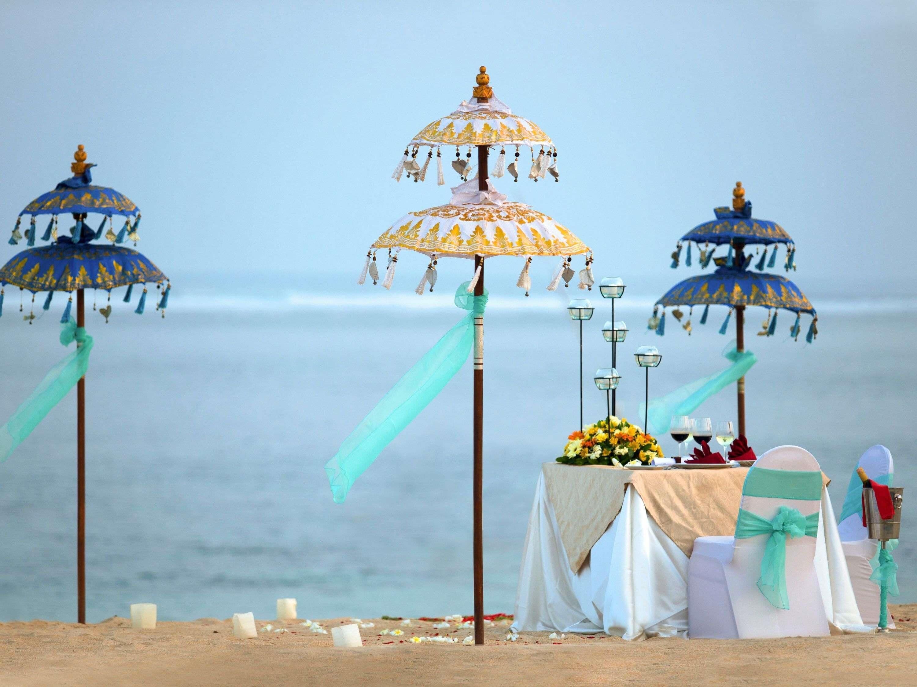 A beach wedding setup with three ornate umbrellas, a table with floral arrangements, and a view of the ocean.