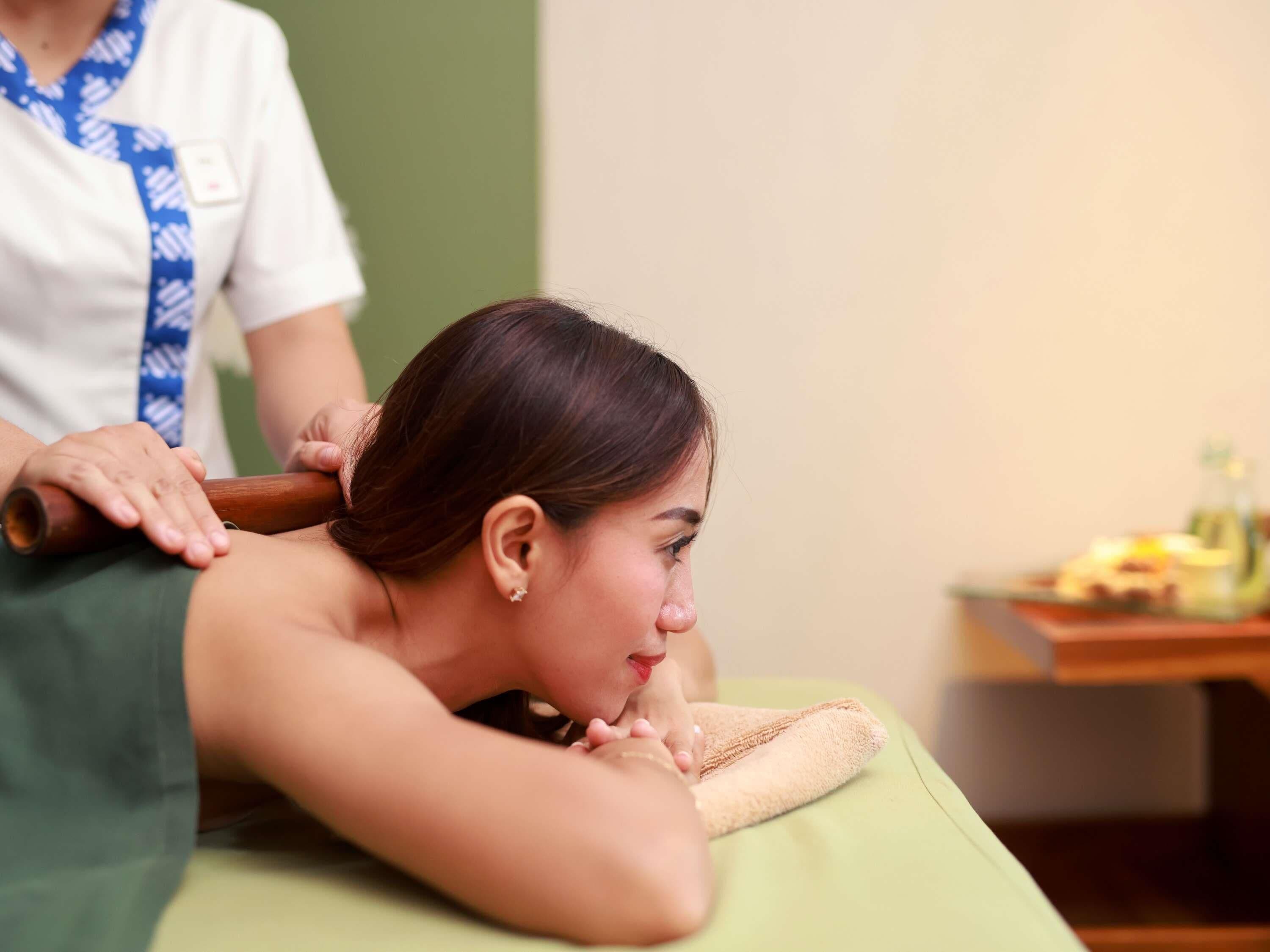 A massage therapy session in progress with a client lying face down on a massage table, a therapist's hands applying pressure to the client's back, and a small table with a bottle and other items in the background.