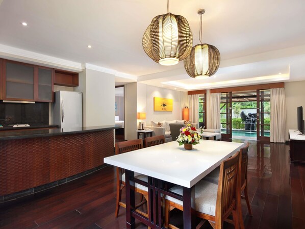 A modern kitchen with a white countertop, wooden cabinets, and a built-in refrigerator. There is a dining area with a white table and wooden chairs, and a large window with curtains. - Novotel Bali Nusa Dua (Nusa Dua)
