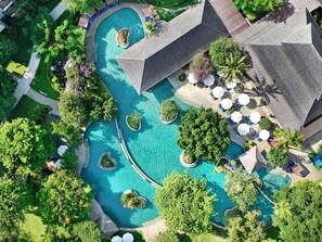An aerial view of a resort with a large, winding pool surrounded by lush greenery and palm trees. There are several lounge areas with umbrellas and seating. - Novotel Bali Nusa Dua (Nusa Dua)