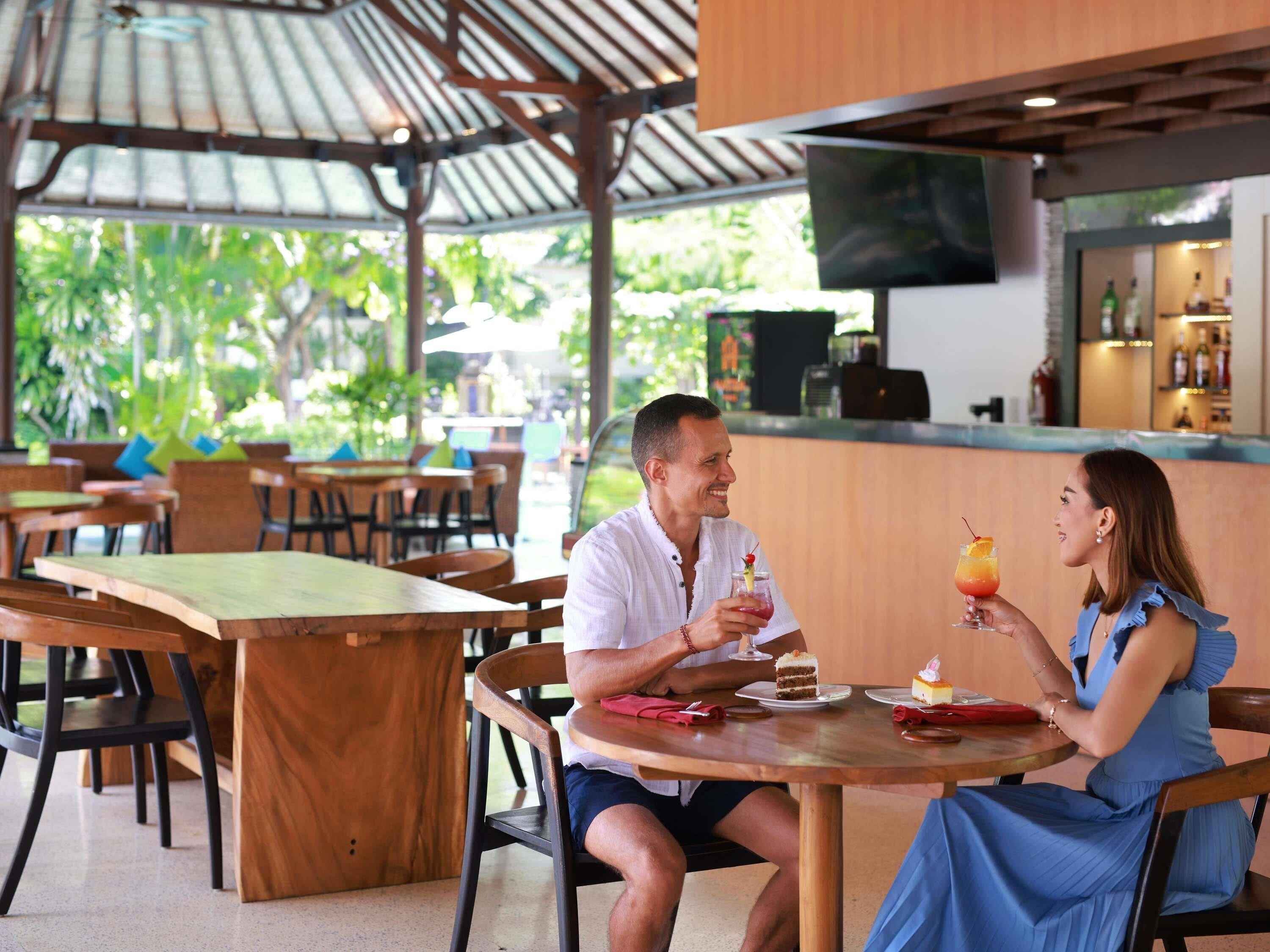 Una pareja cenando en un restaurante al aire libre con mesas y sillas de madera, una zona de bar y vistas a zonas verdes.