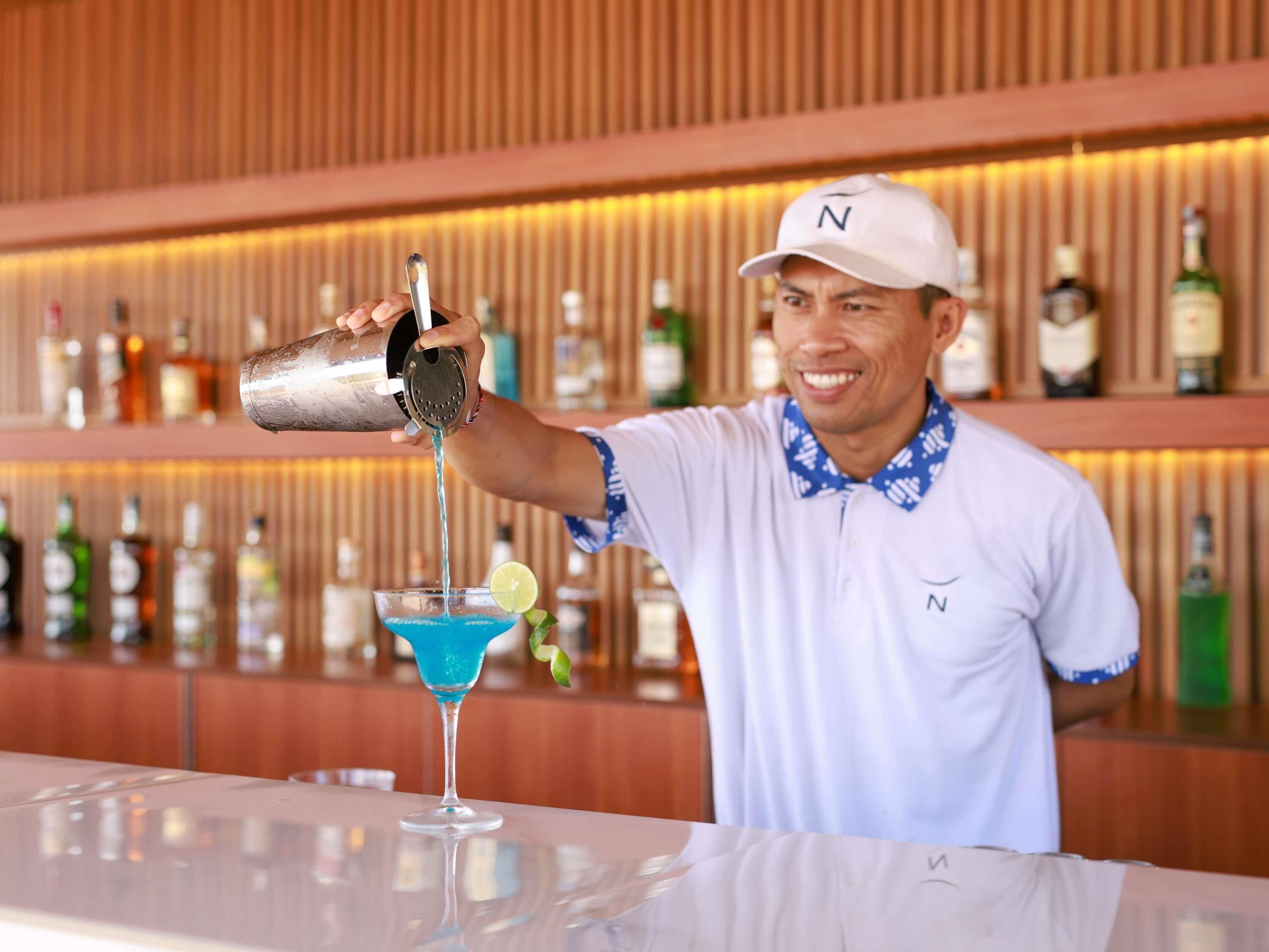 A bartender pouring a blue cocktail into a glass with a lime garnish.