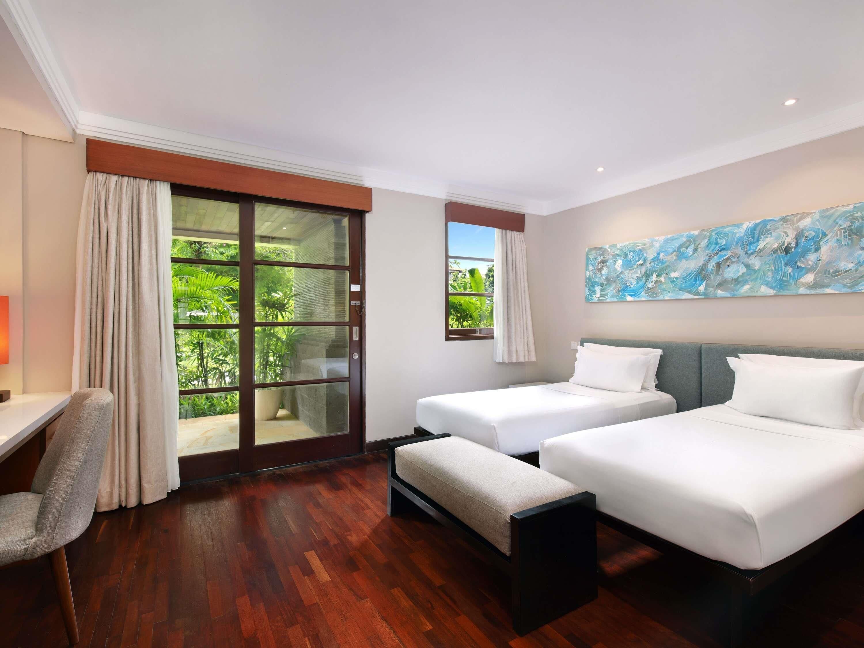 A hotel room with two beds, a desk, a chair, a large window with curtains, and a painting on the wall.