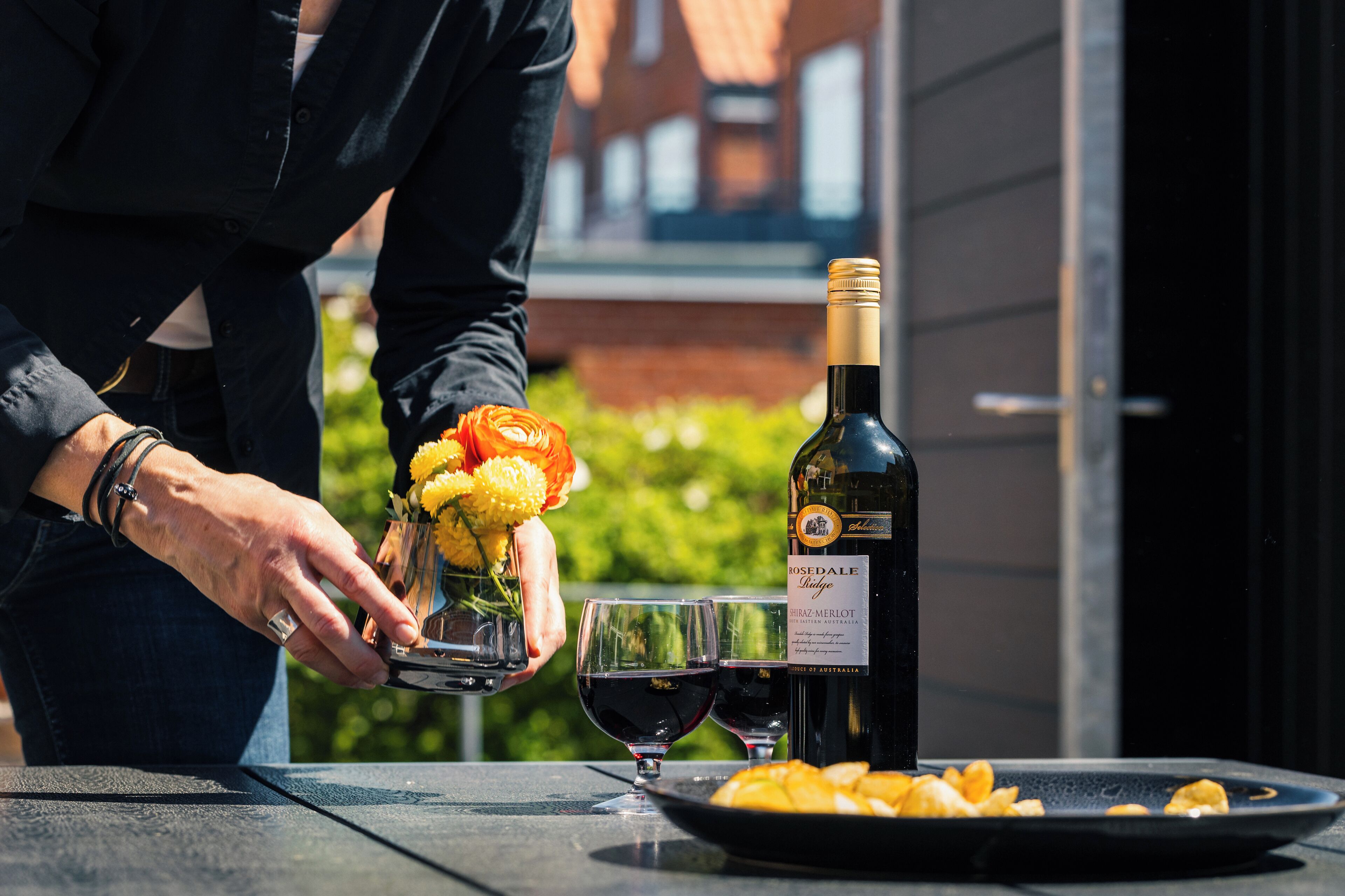Une personne verse du vin dans des verres sur une table en plein air, à côté d’une bouteille de rosé merlot, de fleurs et de quelques amuse-gueules.
