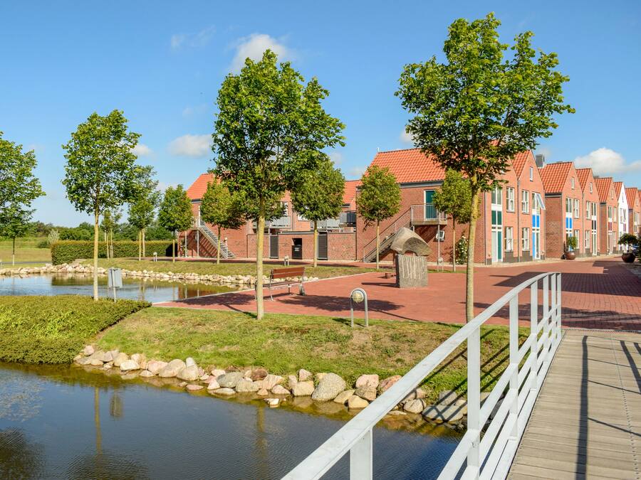 A riverside walkway with brick buildings, trees, and a wooden bridge.