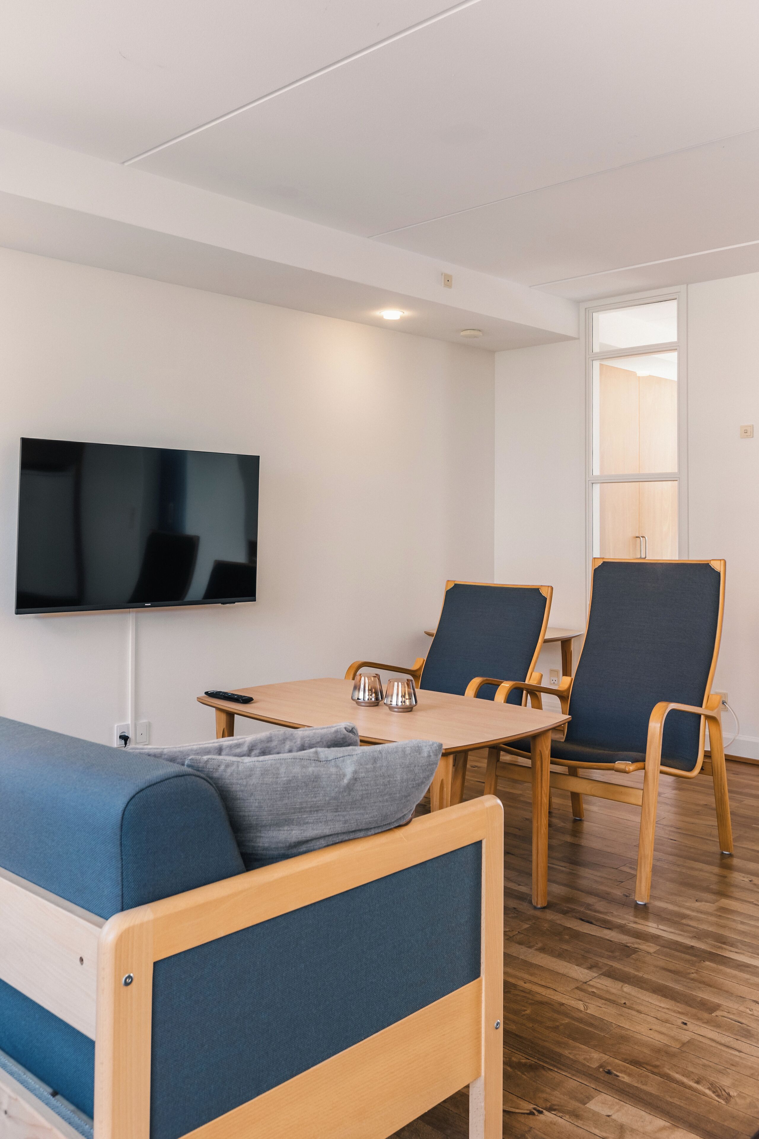 Una camera moderna con un tavolo da pranzo in legno, due sedie, una TV a schermo piatto montata a parete e un divano blu.