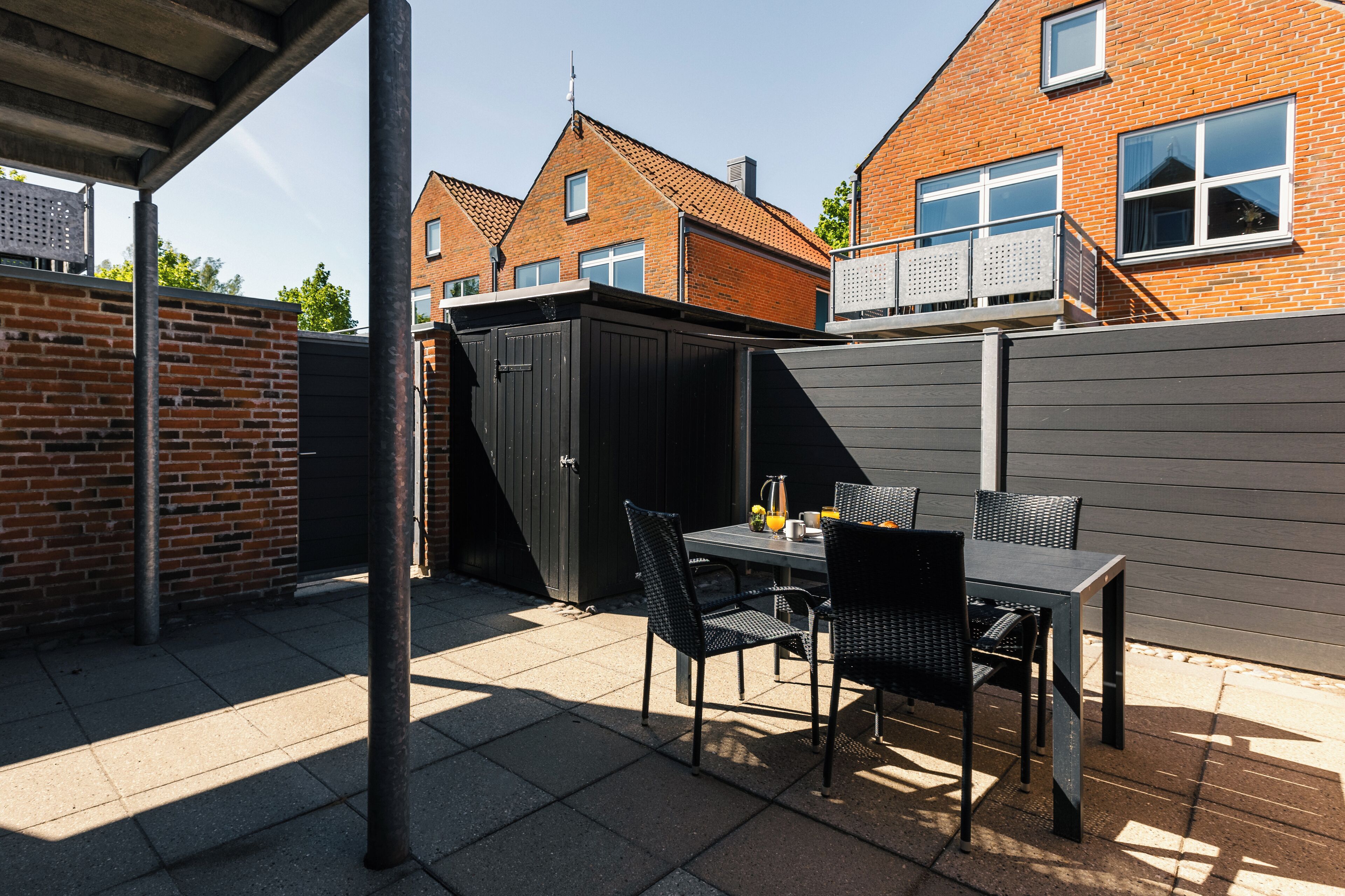Een terras met een tafel, stoelen, een tuinhuisje en een bakstenen muur.