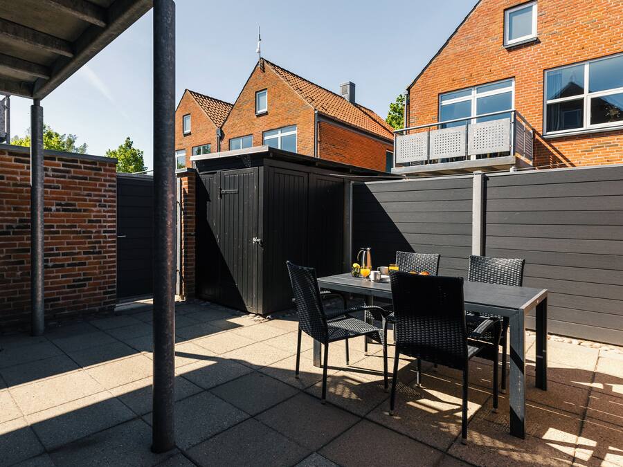 Een terras met een tafel, stoelen, een tuinhuisje en een bakstenen muur.