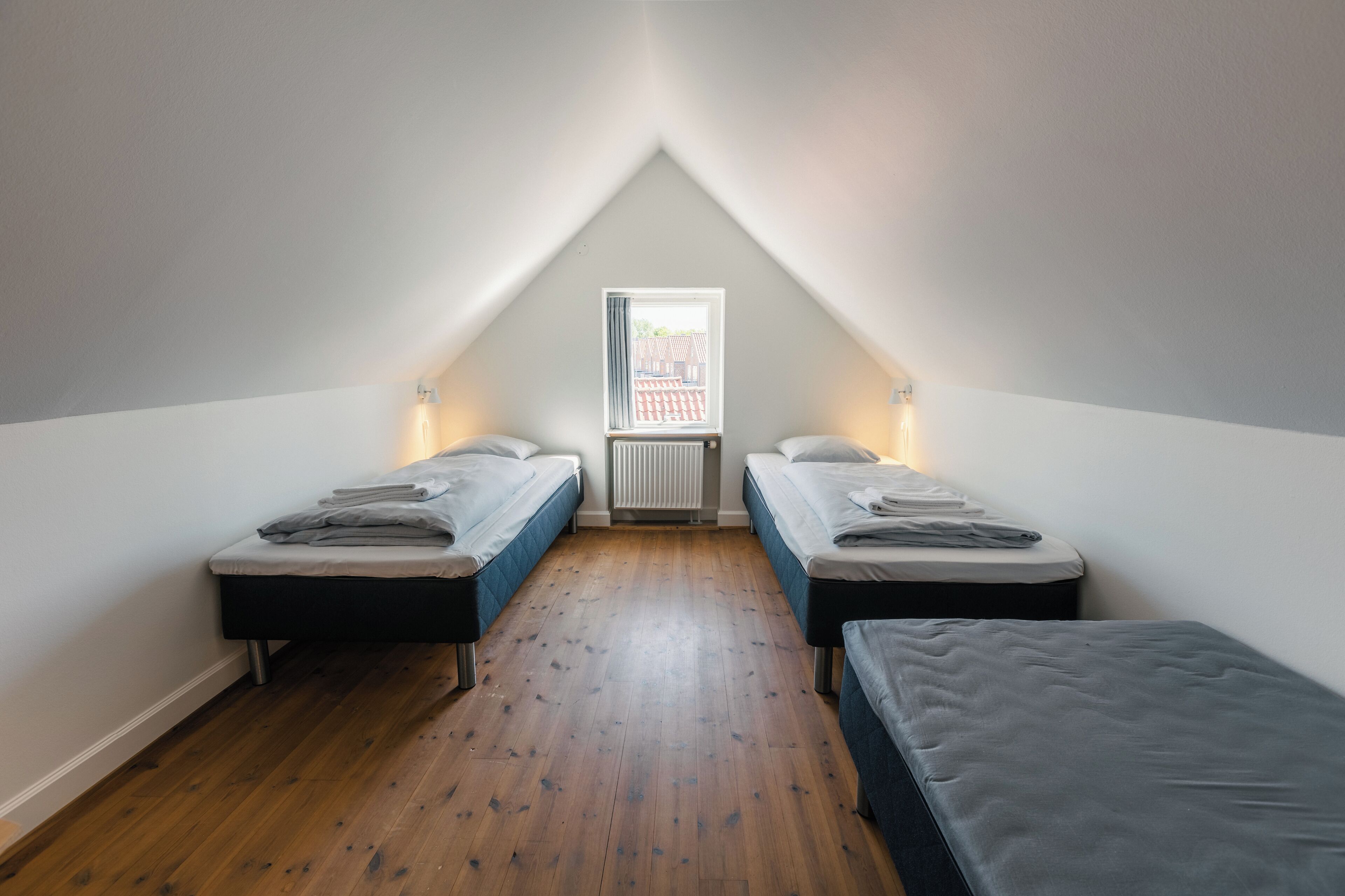 Un dormitorio bajo de techo, pequeño y triangular, con dos camas individuales, un radiador y una ventana que da a un edificio de ladrillo rojo.