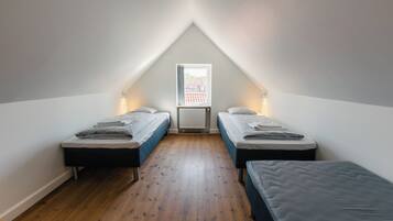 Un dormitorio bajo de techo, pequeño y triangular, con dos camas individuales, un radiador y una ventana que da a un edificio de ladrillo rojo.