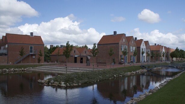 A row of brick houses with a small bridge over a pond, surrounded by greenery and a clear sky.