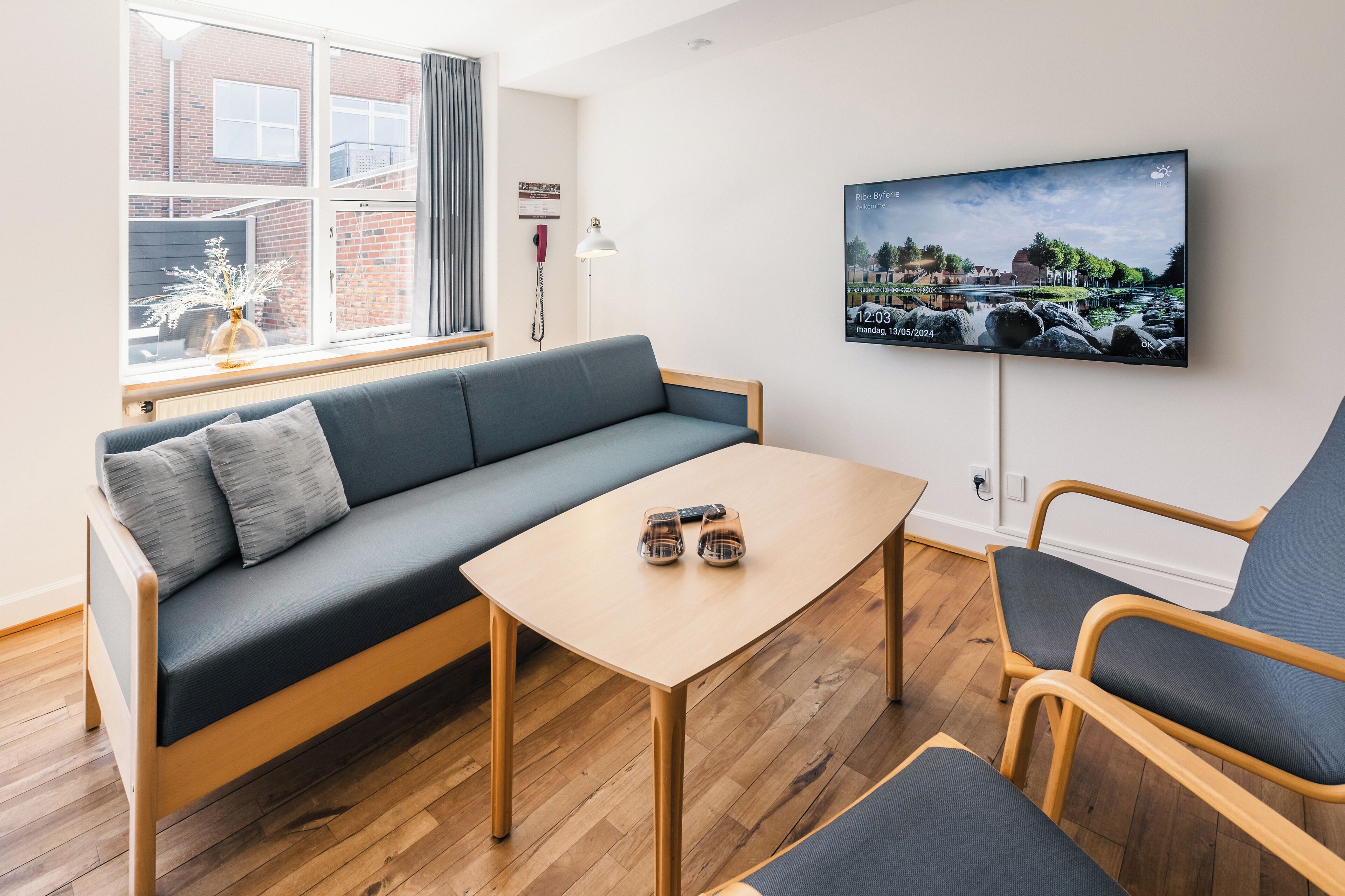 Una sala moderna con un sofá, una mesa de centro de madera y un sillón. Un televisor de pantalla plana está montado en la pared, y una ventana ofrece vistas al exterior.