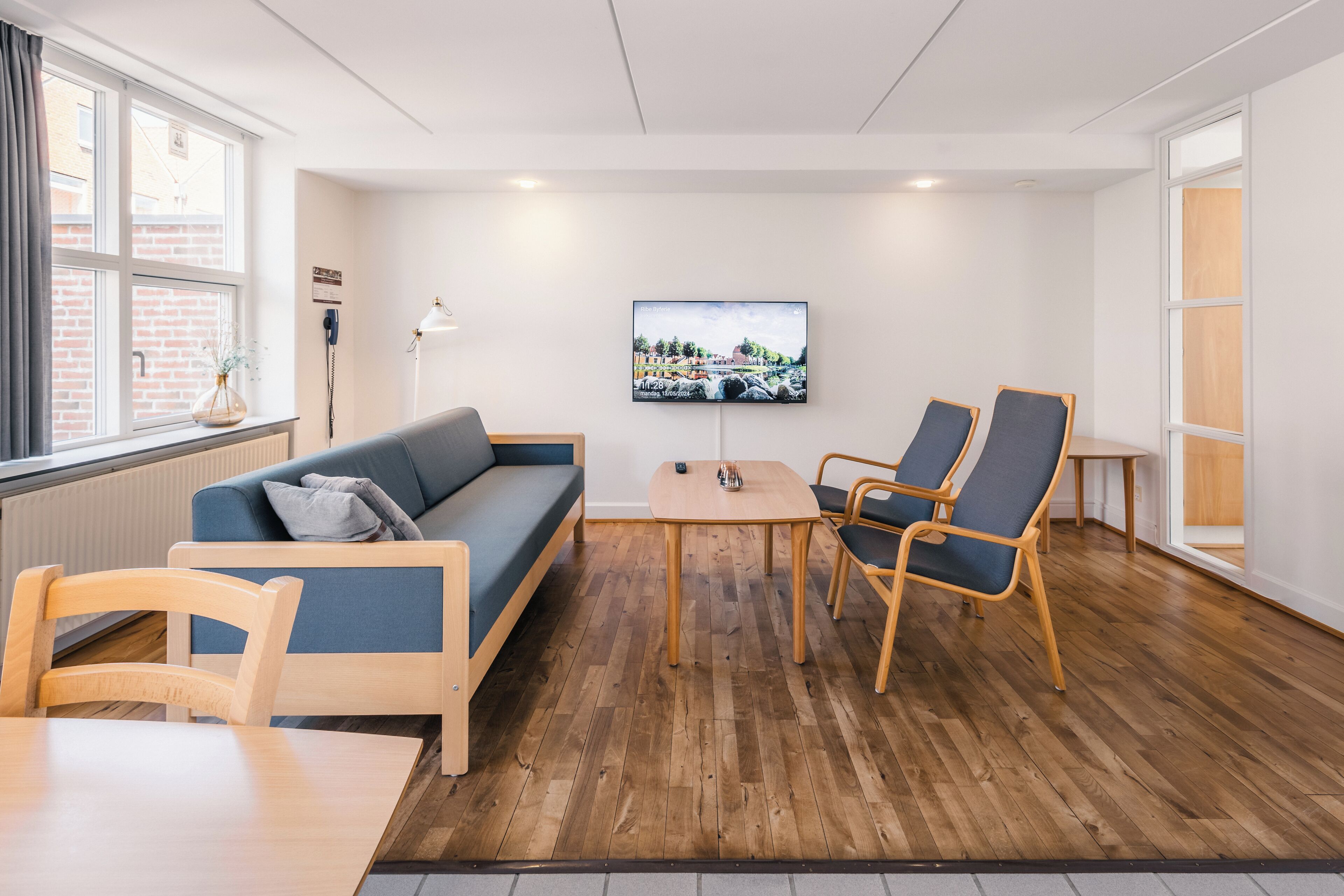 A modern room with a wooden floor, a blue sofa, two armchairs, a small table, and a TV on the wall.