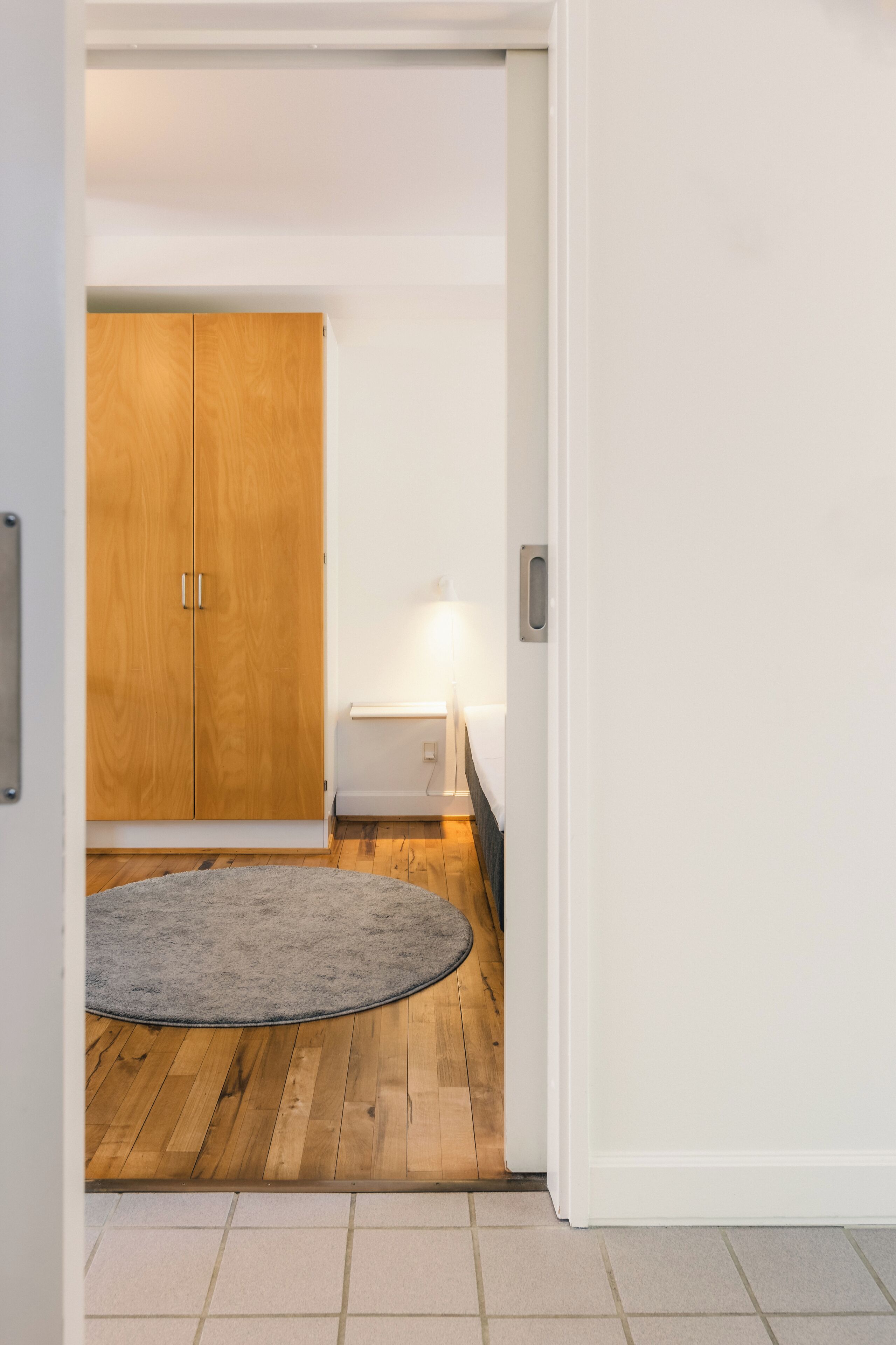 A hallway with a wooden wardrobe, a small table with a lamp, and a grey rug on a wooden floor.