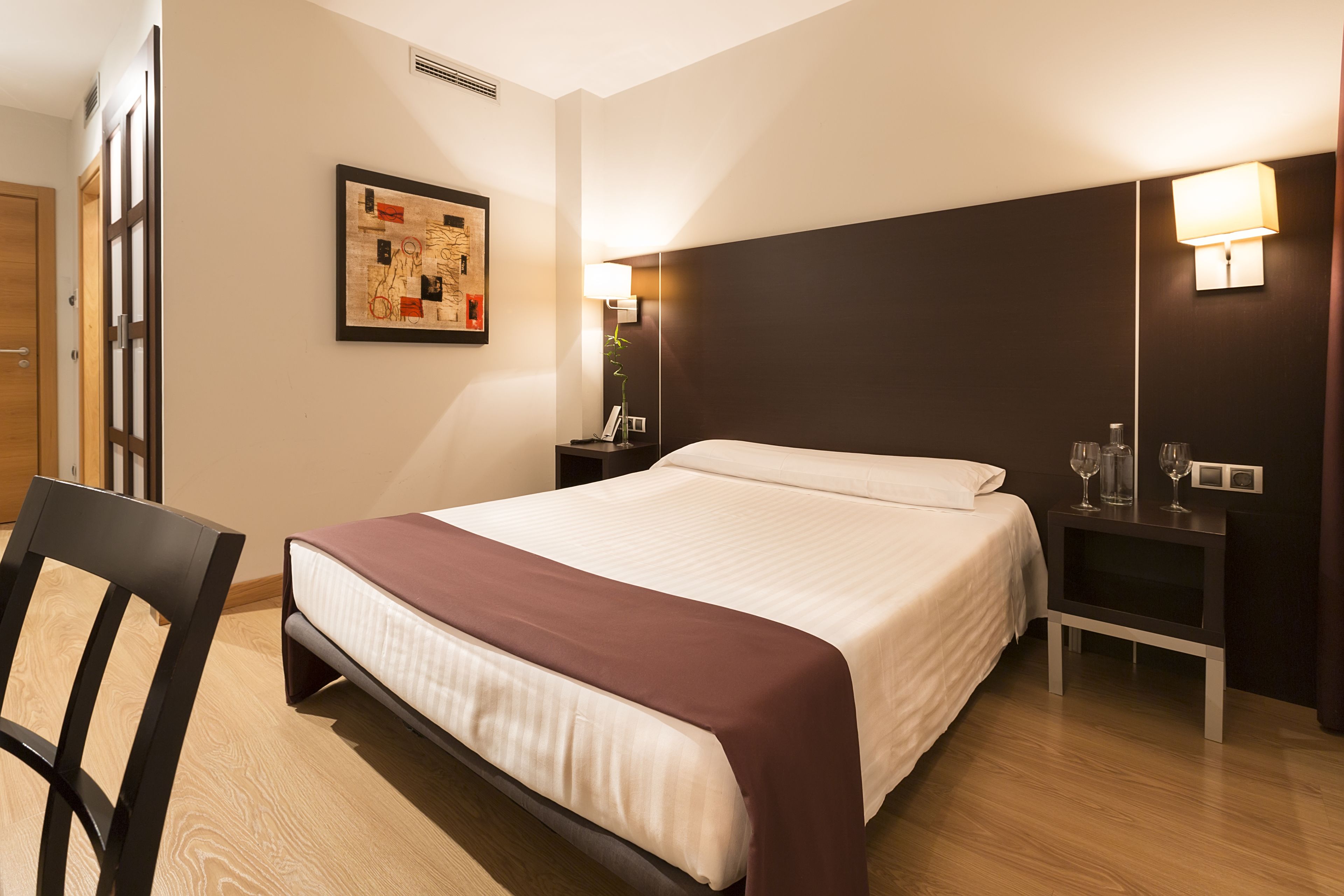 Een hotelkamer met een groot bed, een nachtkastje met een lamp, een stoel en een lijst met een kunstwerk aan de muur.
