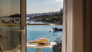 Vista desde un balcón con una mesa que sostiene un sombrero y un vaso de bebida, con vista a una masa de agua con barcos y un horizonte urbano distante.
