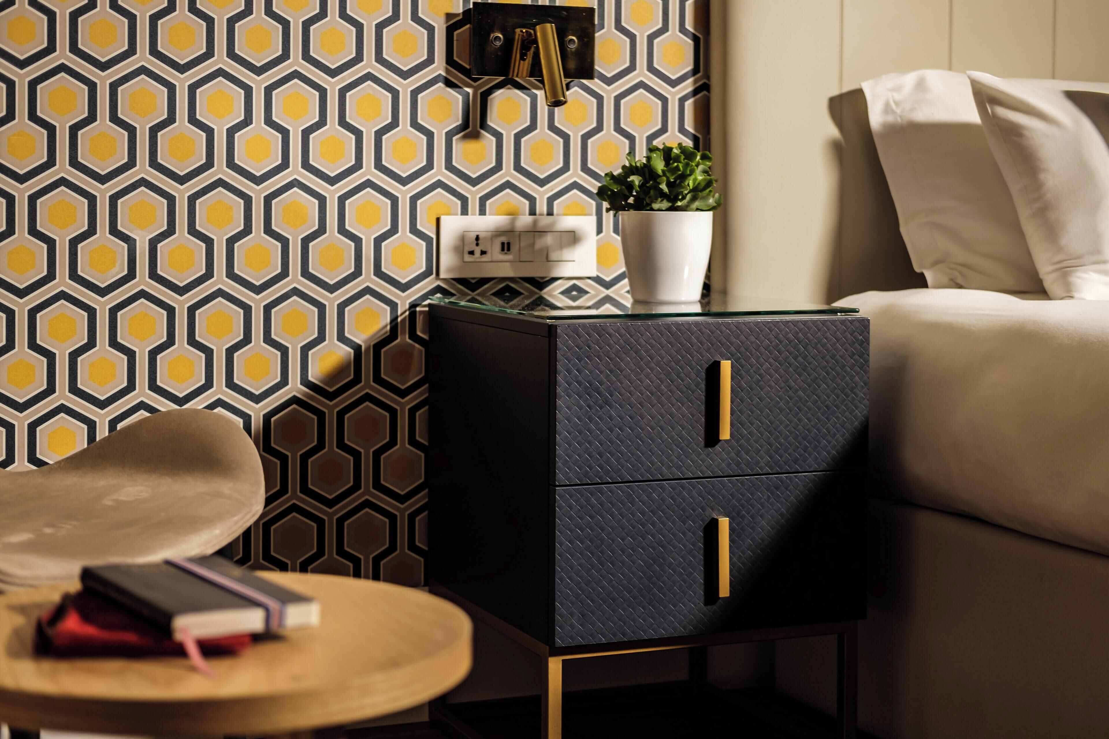 Una habitación moderna con papel tapiz estampado con motivos geométricos, una mesita de noche negra con tiradores dorados, una pequeña mesa redonda y un sofá beige.