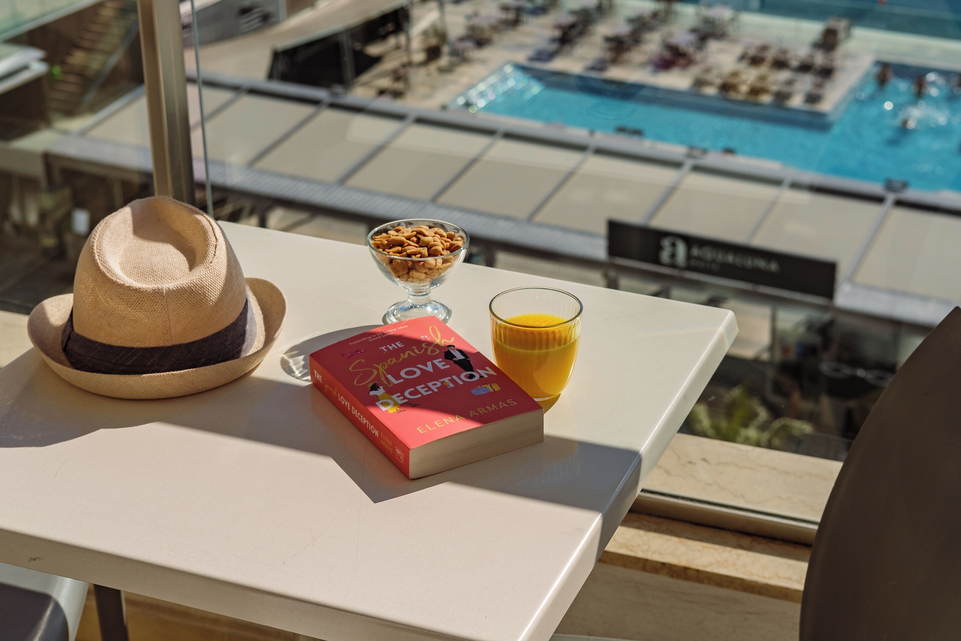Un cappello, un libro intitolato 'The Spanish Love Deception' e un bicchiere di succo d'arancia su un tavolo con vista sulla piscina.