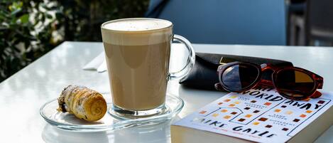 A clear glass coffee cup, a pastry, a pair of sunglasses, and a book titled "World Away" on a white table outdoors.