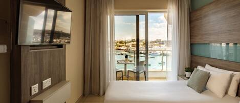 Habitación de hotel con una cama grande, un televisor montado en la pared, un balcón con vistas al puerto deportivo y una pequeña mesa con una planta.