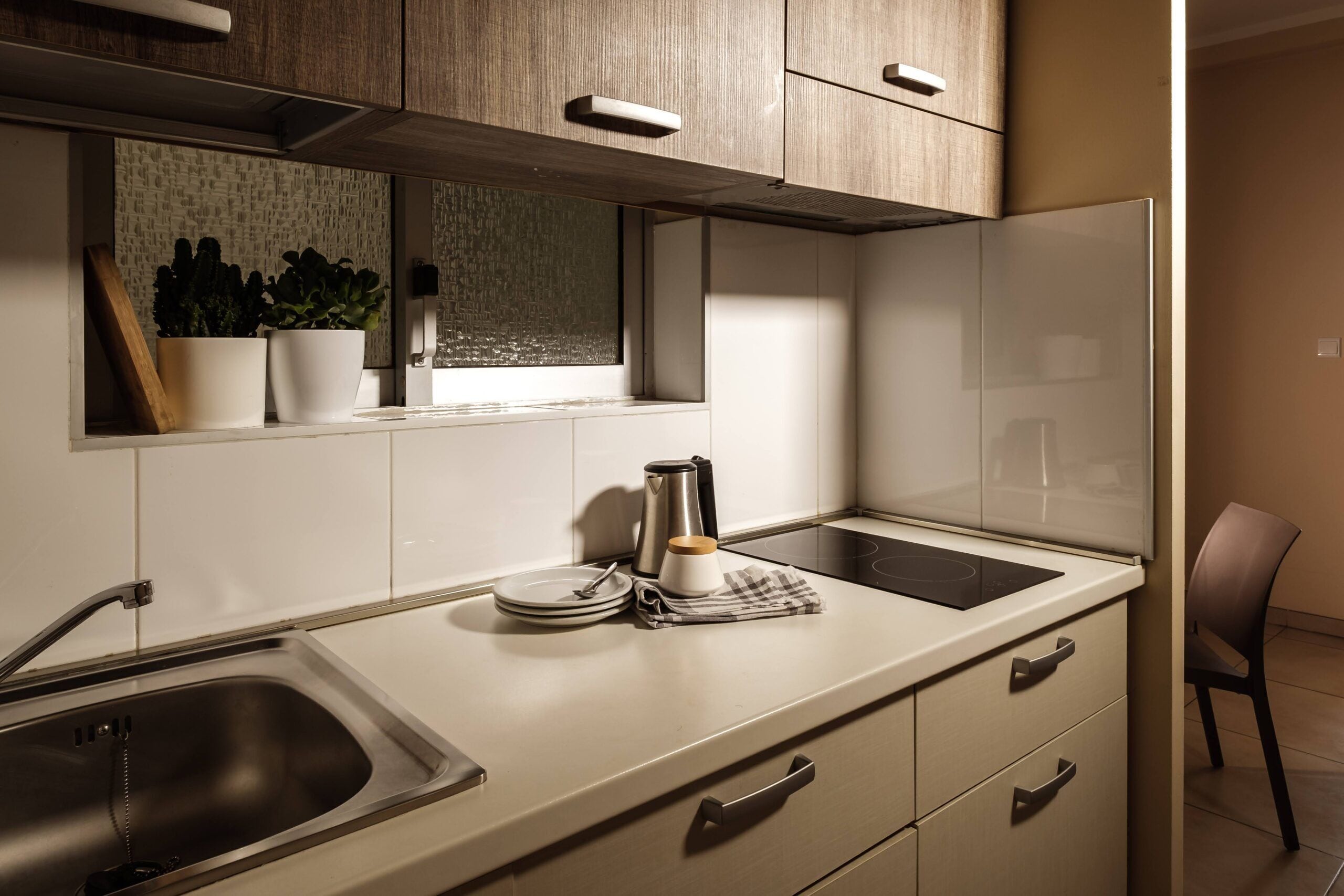 A modern kitchen with a stainless steel sink, a window with plants, a kettle, and a chair.