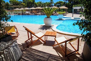 Un área junto a la piscina con sillones de descanso y sombrillas, rodeada de abundante vegetación.