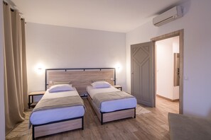 Una habitación de hotel con dos camas individuales, cabecero de madera, mesitas de noche y una puerta que da a otra habitación.