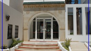 A building with a traditional architectural style, featuring a central arched entrance, a tiled roof, and a staircase leading to the entrance. - Hotel Emira (Hammamet)