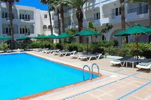 A swimming pool with lounge chairs and umbrellas, surrounded by palm trees and a multi-story building with balconies. - Hotel Emira (Hammamet)