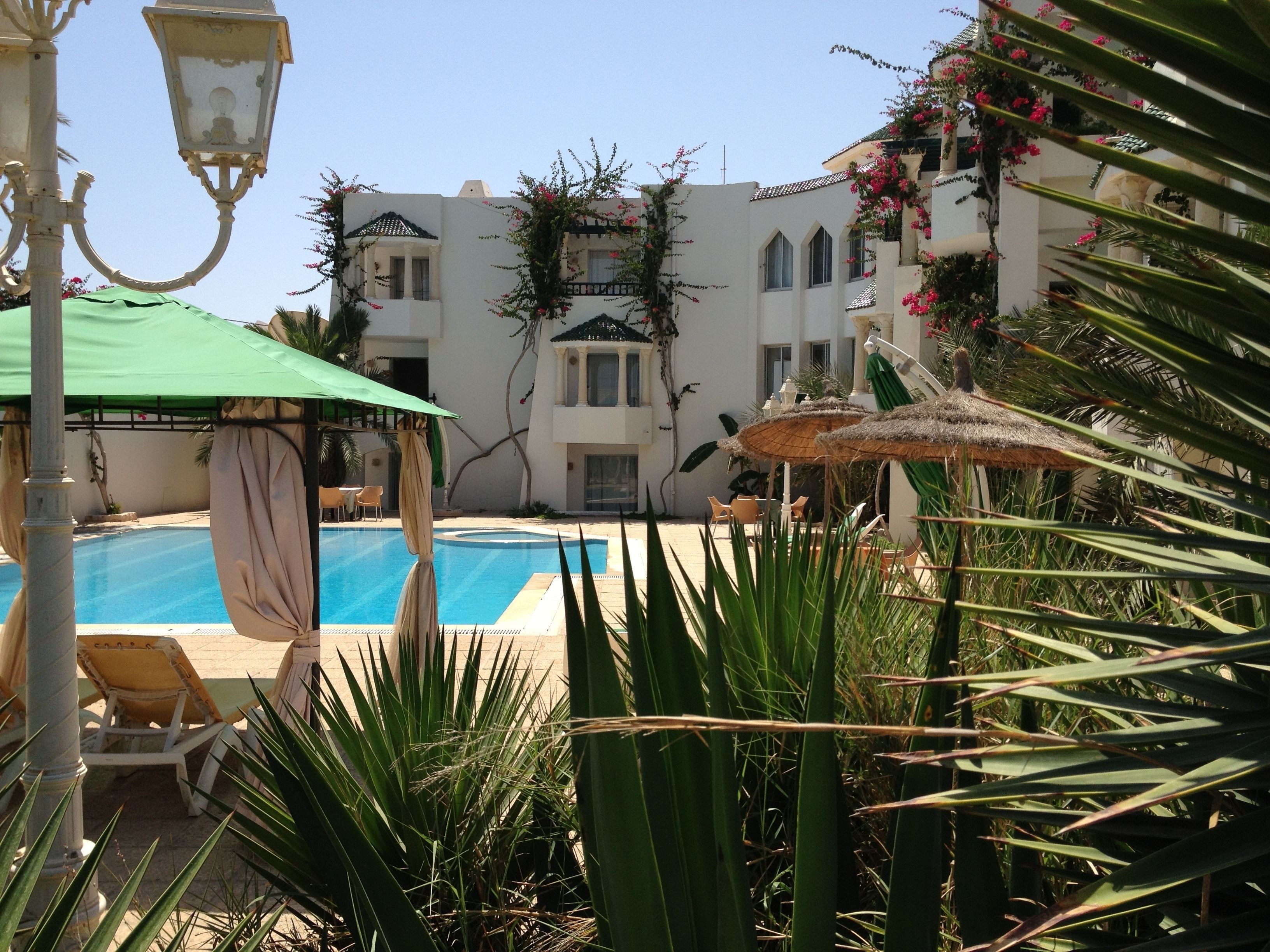 Zona de piscina con sillones de descanso y sombrillas, rodeada de vegetación y un edificio con varias ventanas y balcones.