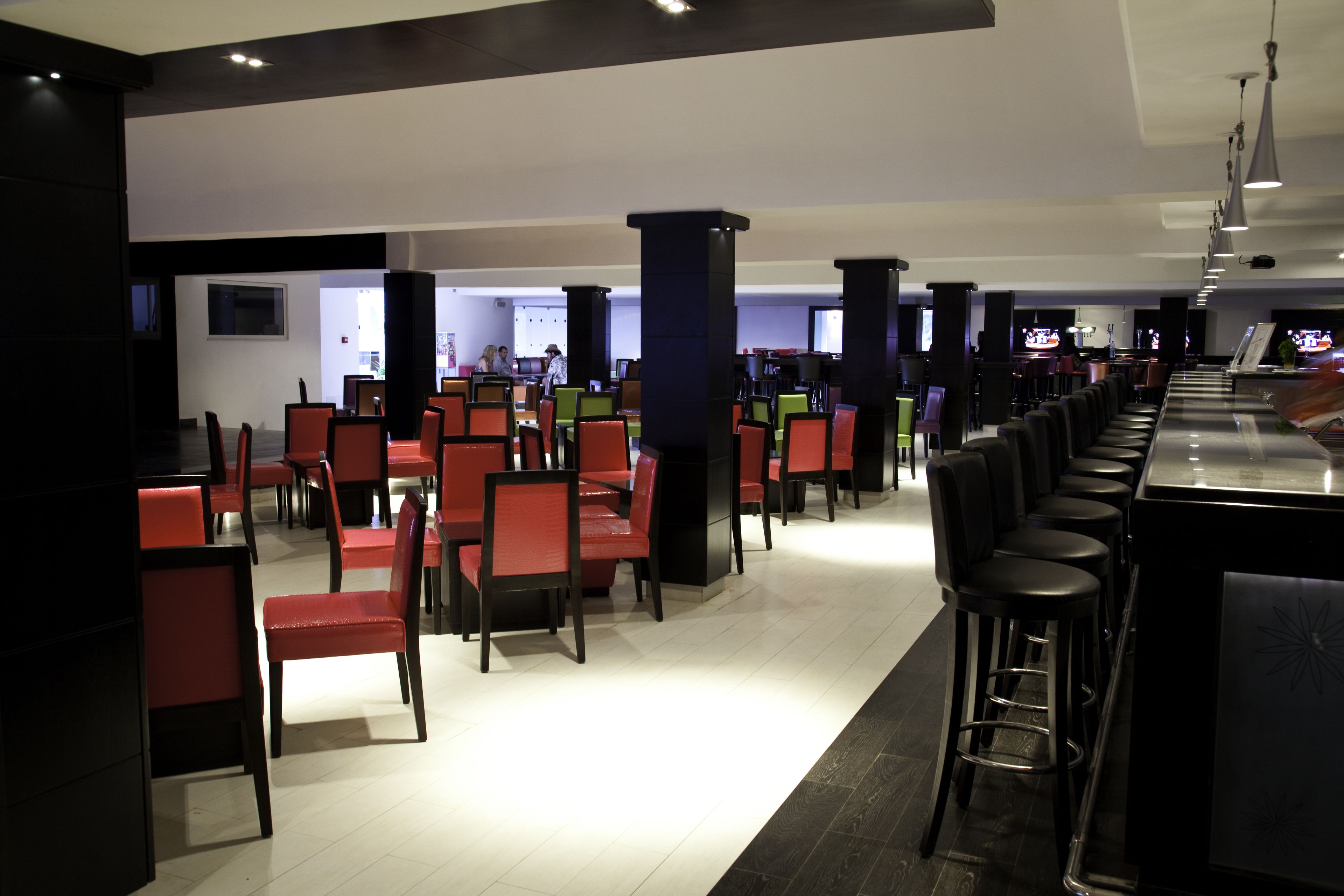 Un restaurant moderne avec des chaises rouges et noires, un coin bar avec des tabourets et un sol carrelé, propre et élégant.