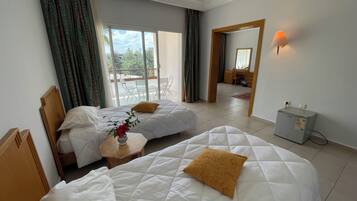 A hotel room with two beds, a small table with a vase, a desk with a lamp, and a view of the outside balcony.