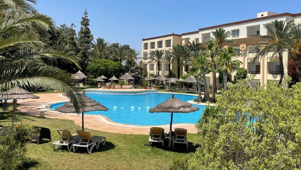 A resort pool area with lounge chairs and umbrellas, surrounded by palm trees and a building with balconies.