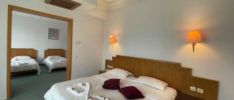 Habitación de hotel con una cama grande, dos mesitas de noche, cabecera de madera, lámpara de pared y una puerta que da a otra habitación.