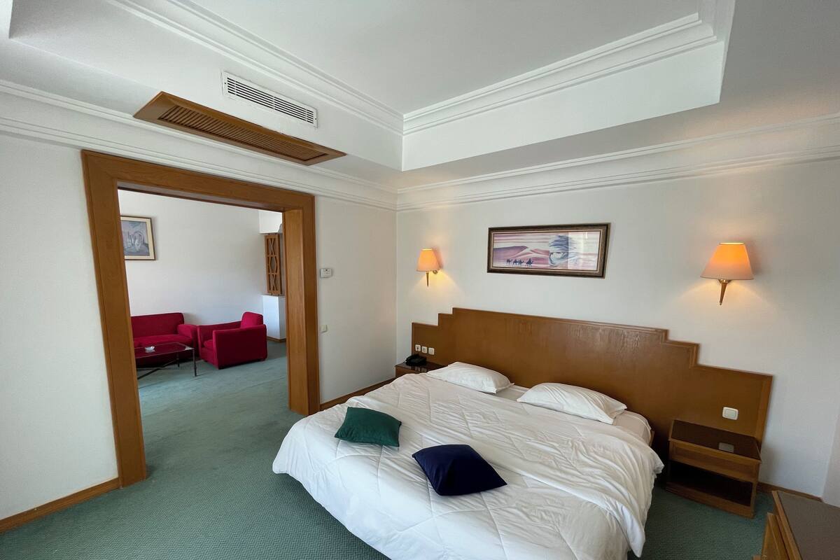 Ein Hotelzimmer mit einem großen Bett, zwei Nachttischen, einem Schrank, einem roten Sessel, einer Holztür und einem gerahmten Bild an der Wand.