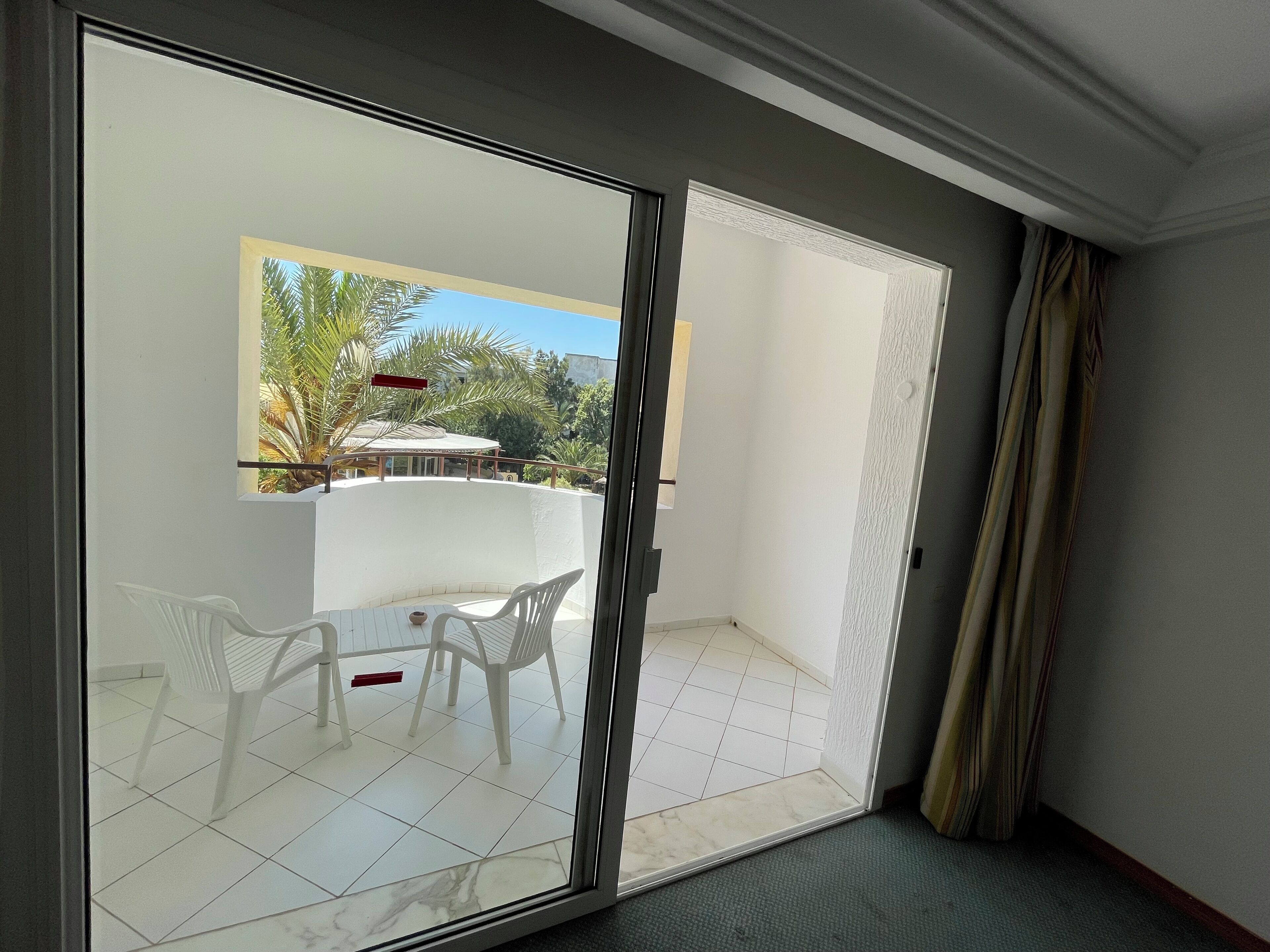 A room with a sliding glass door leading to an outdoor patio with a table and chairs, a view of greenery, and a white tiled floor.